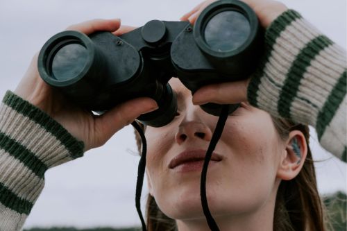 A woman uses binoculars outdoors, engaging in nature observation on a cloudy day.